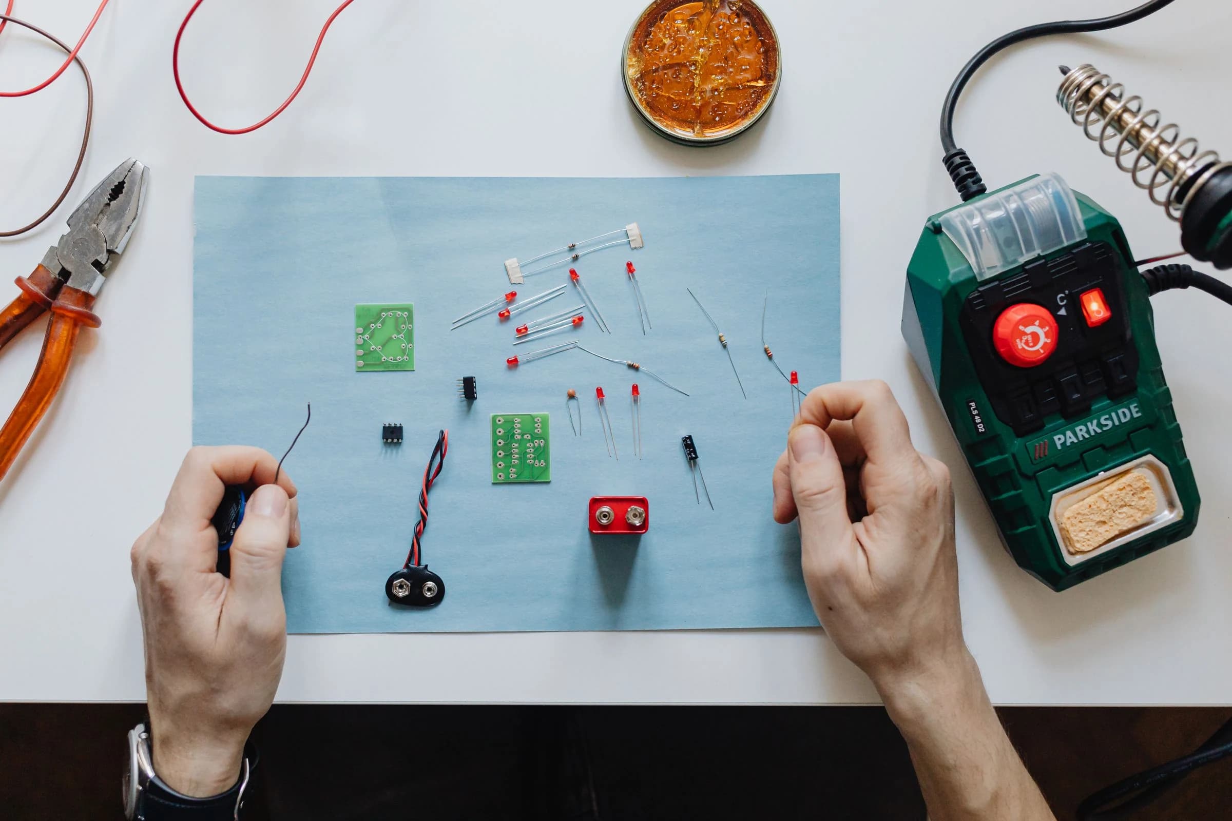 An overhead view of hands assembling electronics at a workbench with circuit boards, components and soldering tools.