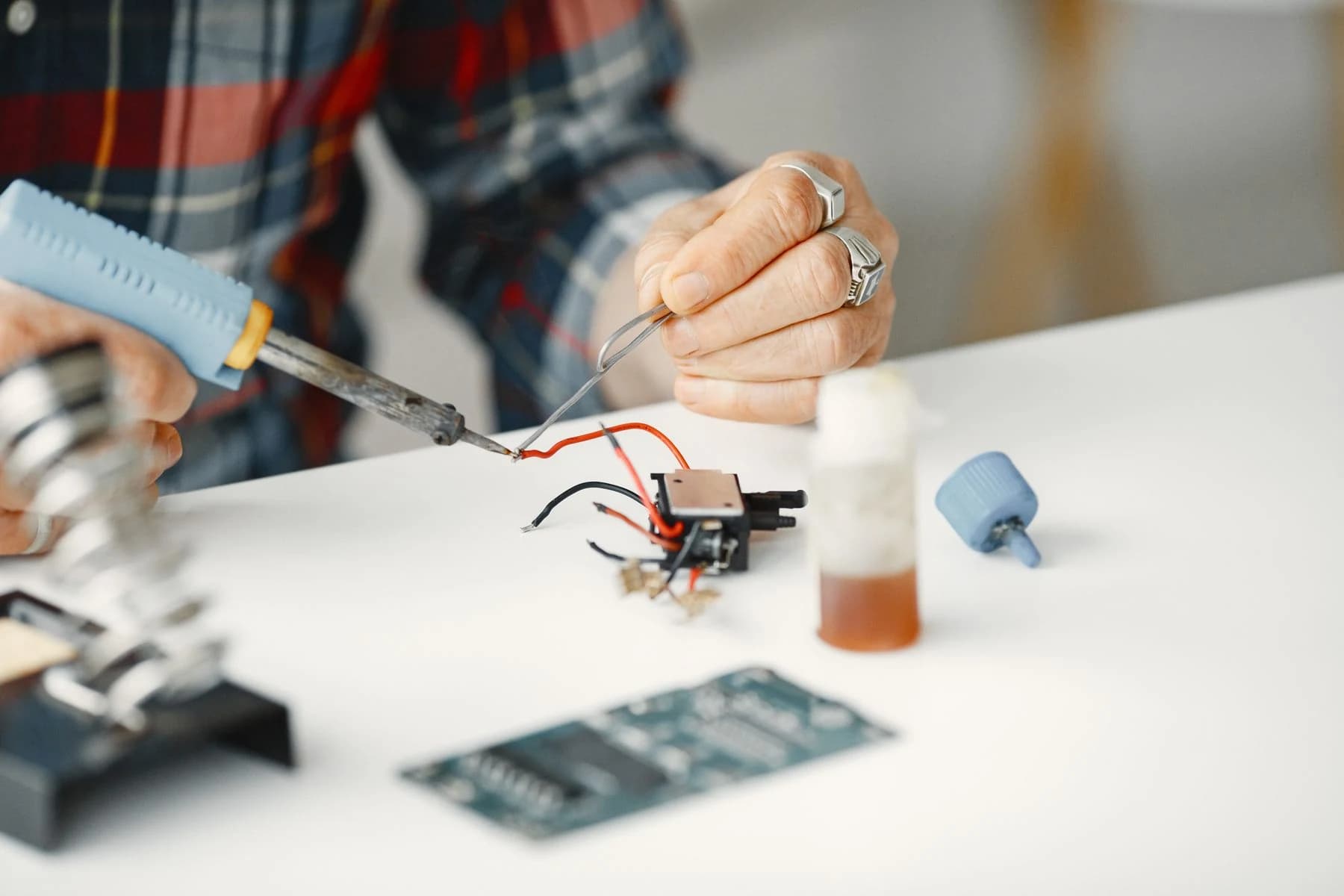 A close-up of hands soldering wires to an electronic component on a white worktable.